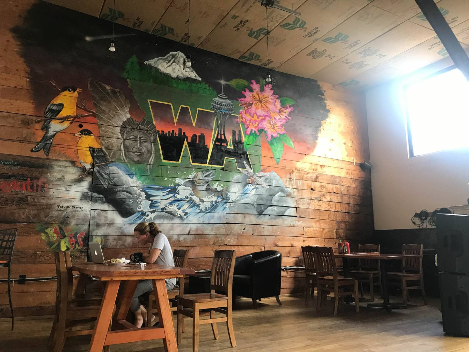 A cafe setting with wooden tables and chairs, and a wooden wall with a giant colourful mural reading “WA” with some icons of the state of Washington in USA. A woman sits with a laptop at one of the tables, deep in focus.