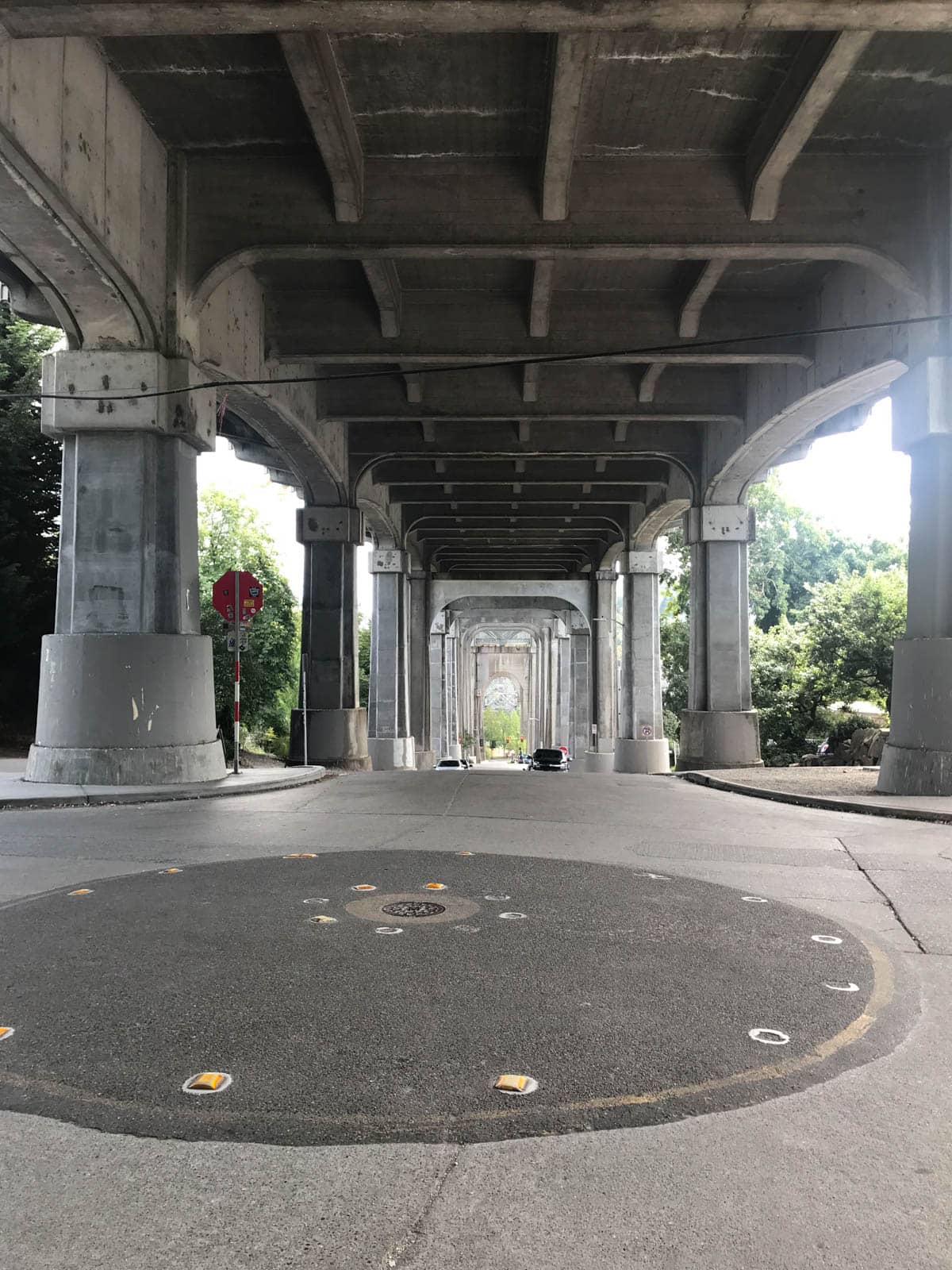 A view of the street under a bridge, across the length of the bridge. The bridge’s pillars are in view, and some cars can be seen in the distance parked by the sides of the street.