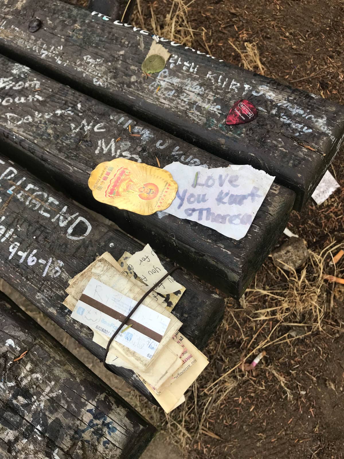 A close up of a bench with some writing painted on it in white, “We miss you”, “legend”, and some handwritten notes tied to the bench, one reading “Love you Kurt”.