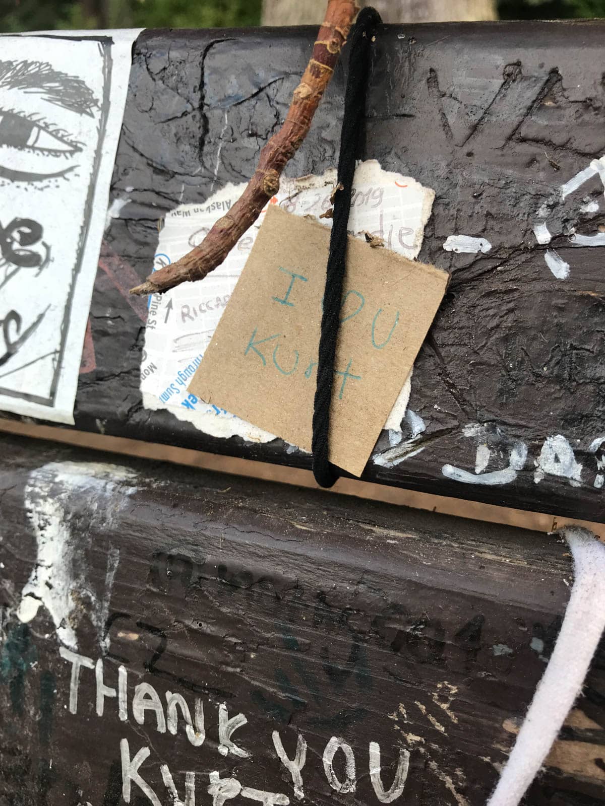 A close up of an old bench with words painted on it reading “thank you Kurt”. Some notes and messages on paper are tied to the bench.