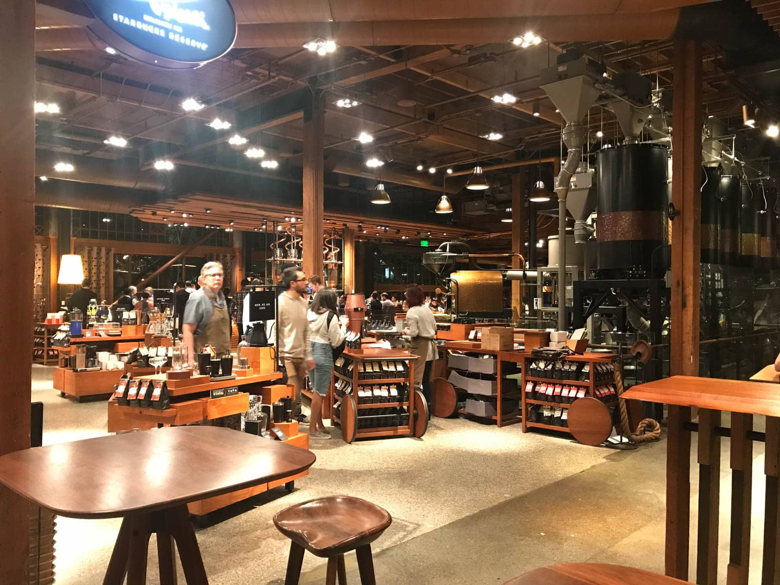 The inside of a cafe with multiple wooden shelves and tables. The shelves have products available for purchase including mugs and coffee. The space is artificially lit, and it’s dark outside.