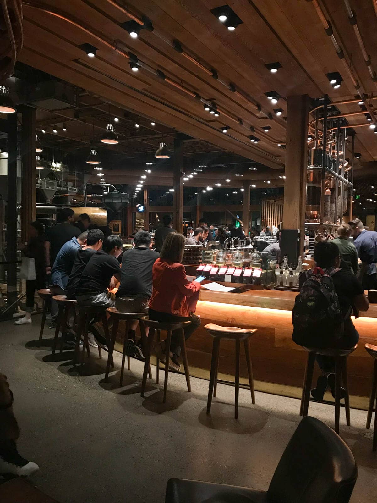 People sitting on high chairs at a high table inside of a cafe. Someone can be seeing working behind the bar. The place is dimly lit with artificial lighting.