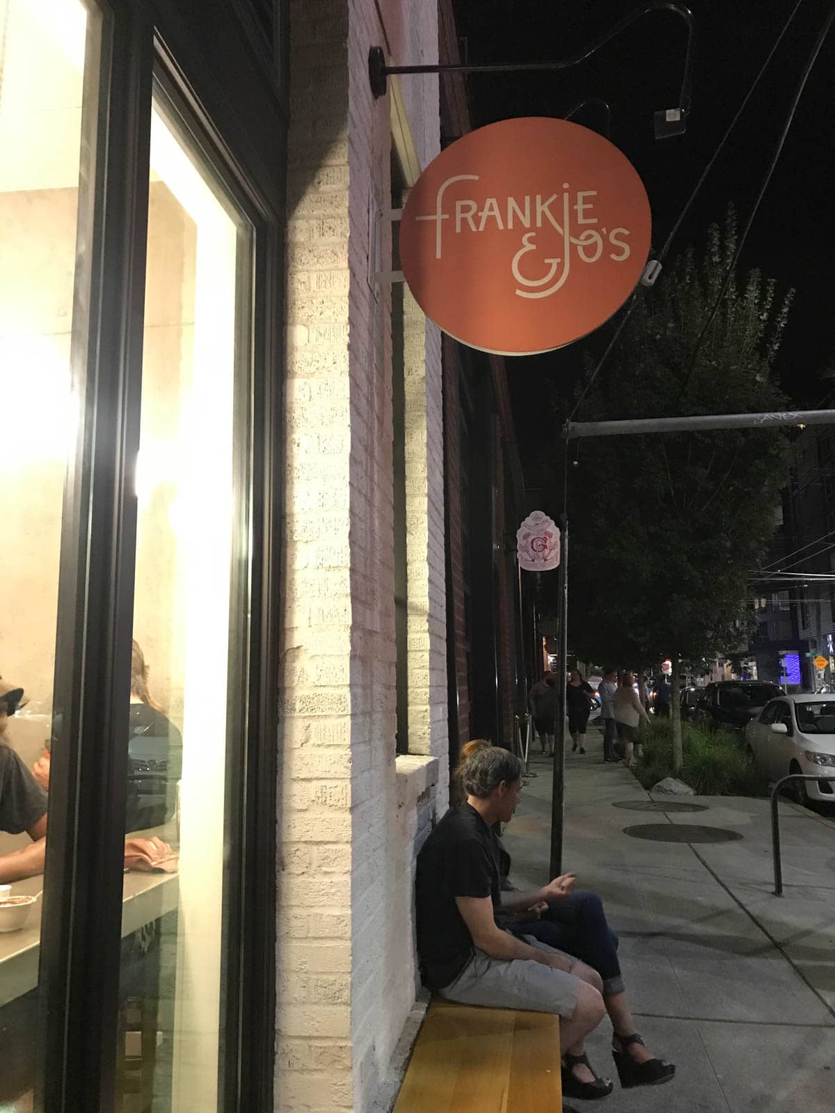 A side view of the outside of an ice cream parlour with a round pink sign reading â€œFrankie & Joâ€™sâ€�. Two people sit on a bench outside the parlour, and some people can be seen sitting inside though the glass window.