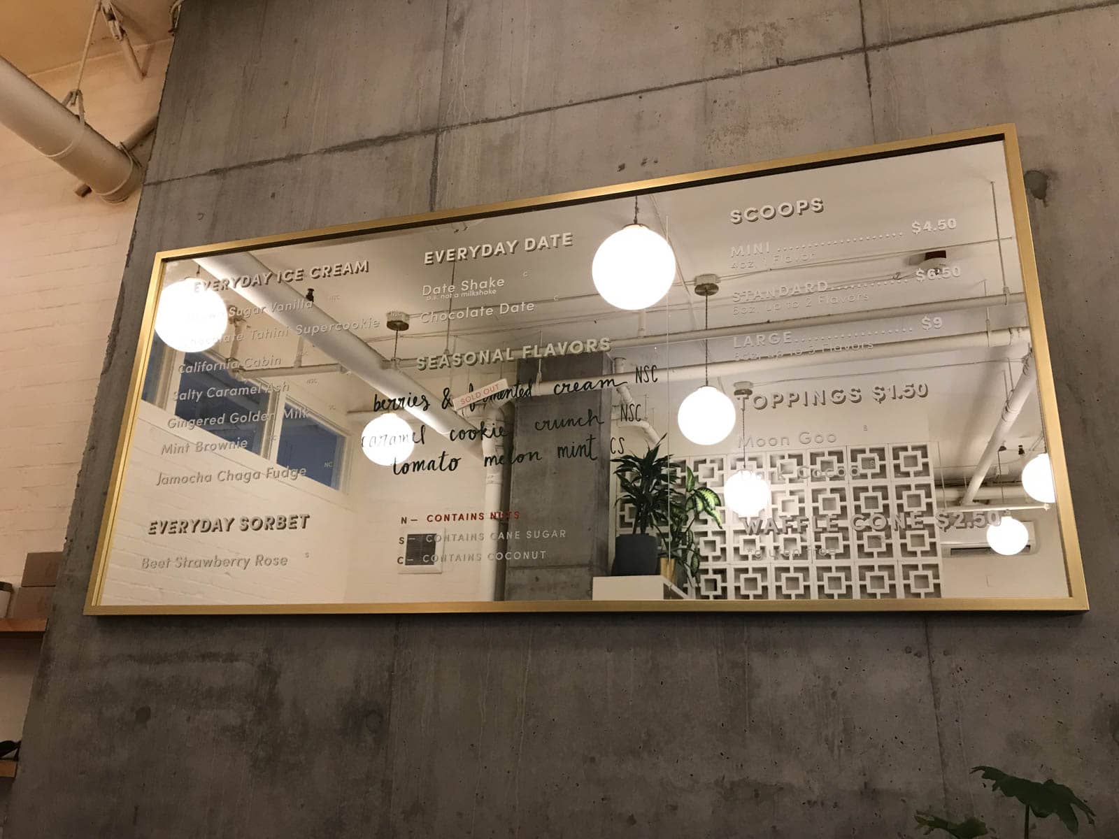 An ice cream menu indoors, printed on a mirrored surface. In the mirror’s reflection, big globe lights hanging from the ceiling can be seen.