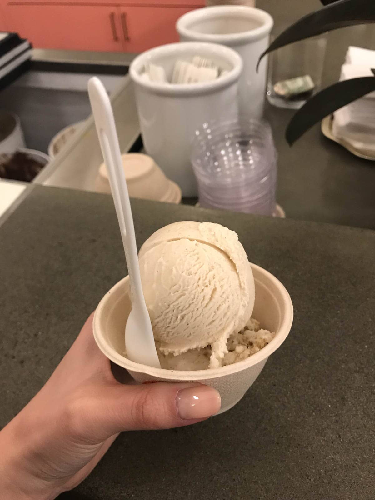 A close shot of a scoop of ice cream in a small cardboard cup. A plastic spoon sticks out of the cup, which is held in someone’s hand.