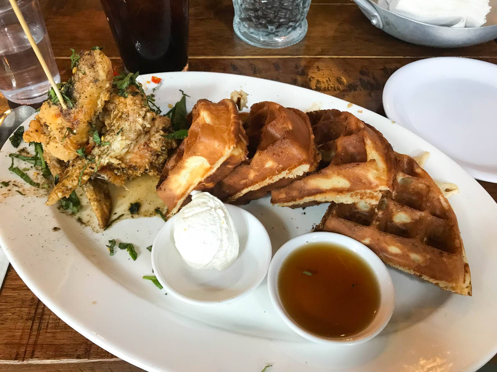 A white oval plate with two small saucers of ice cream and sweet sauce, and a round waffle cut into quarters and a standing shower of chicken wings, garnished with herbs.