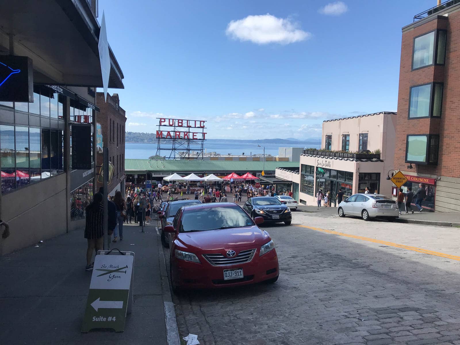 The side of a street heading down towards the entrance of a covered indoor market by the water. Some stalls with umbrella covers can be seen in front of the entrance. A large sign above the roof of the market reads “Public market”.