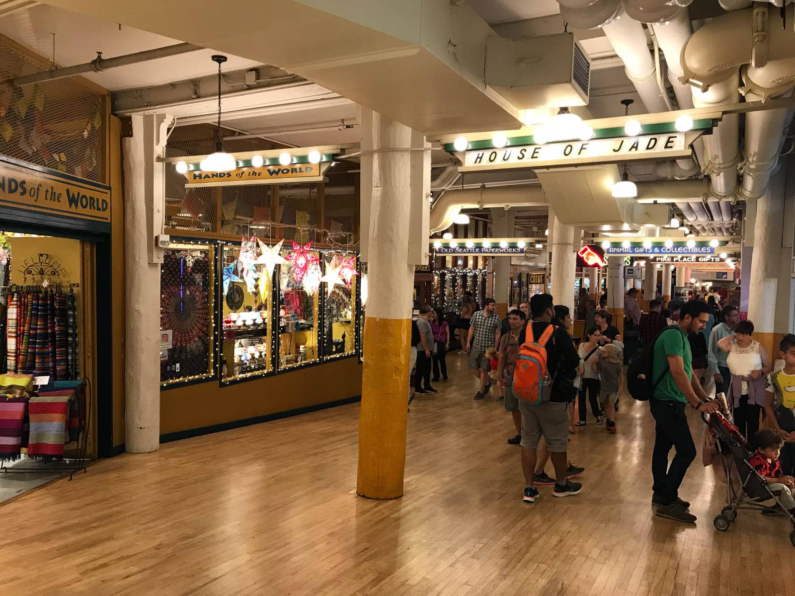 The inside of a building, dimly lit, with some glass shopfronts visible. A sign towards the ceiling reads “House of jade”. Some people can be seen, with backpacks, and one person with a pram and children, in the common area in front of the storefronts.