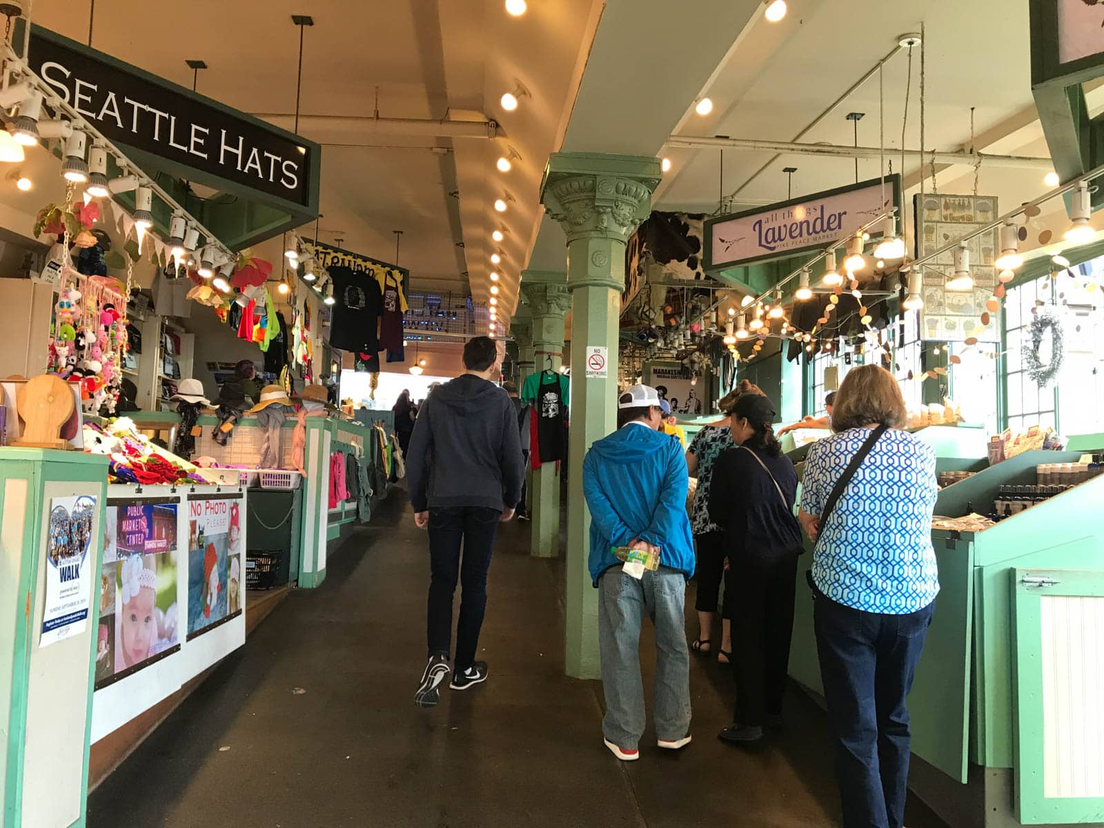 Some people walking up a gradually sloping path between the stalls of an indoor market. One sign reads “Seattle Hats” and another reads “Lavender”.