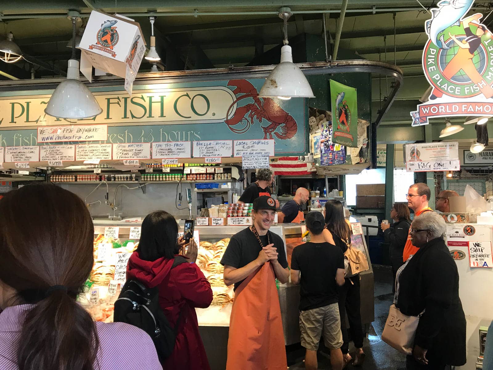 The front of a fish shop inside an indoor market. Some of the seafood can be seen. There are several people gathered around the shop.