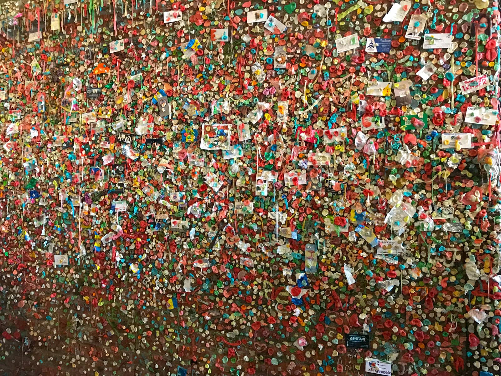 A brick wall littered with various pieces of paper, some resembling business cards and photographs, stuck to the wall with an infinite amount of small pieces of brightly coloured, masticated chewing gum.