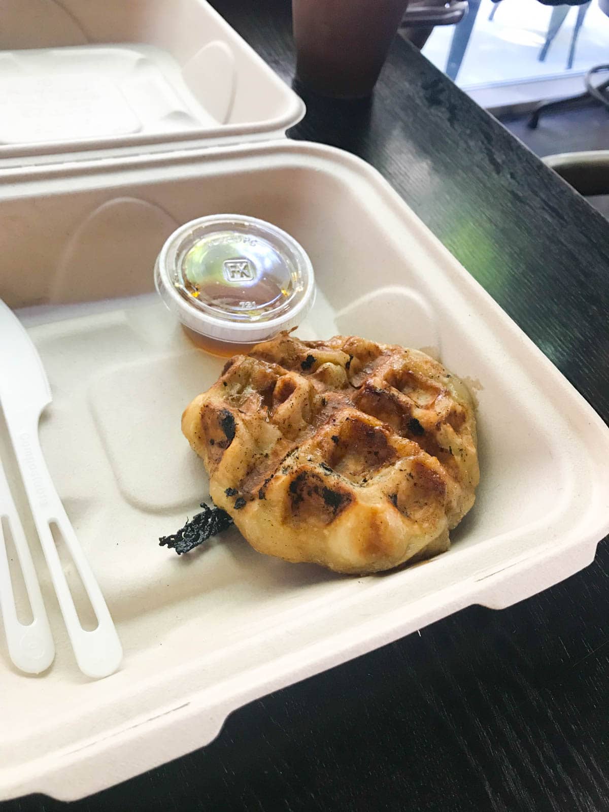 A small waffle in a white styrofoam box with some sweet syrup in a small cup next to it.