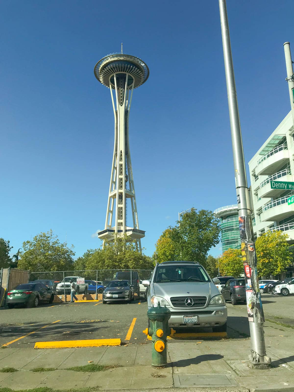 A tall structure with a round observation neck up top. In the foreground is an open car park with some cars parked in it.