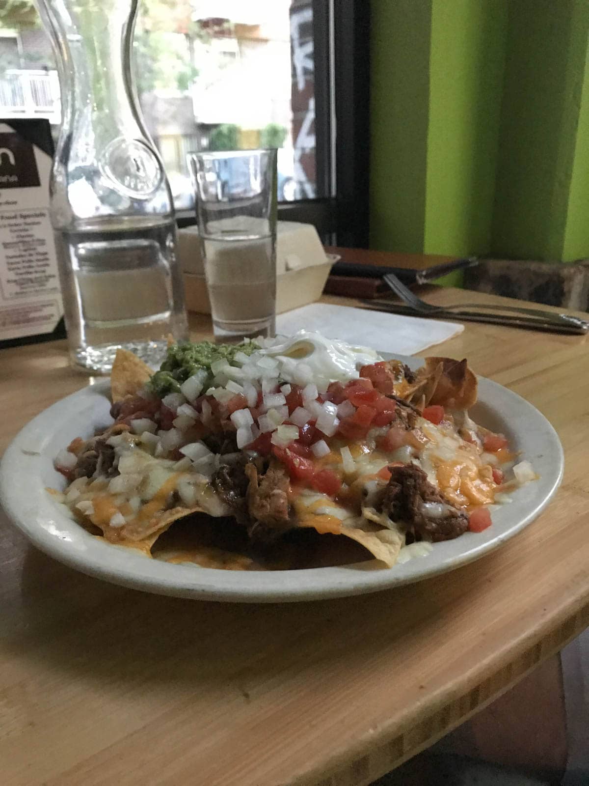 A plate of nachos with toppings of pico de gallo, avocado and onion, sitting on a wooden high table.