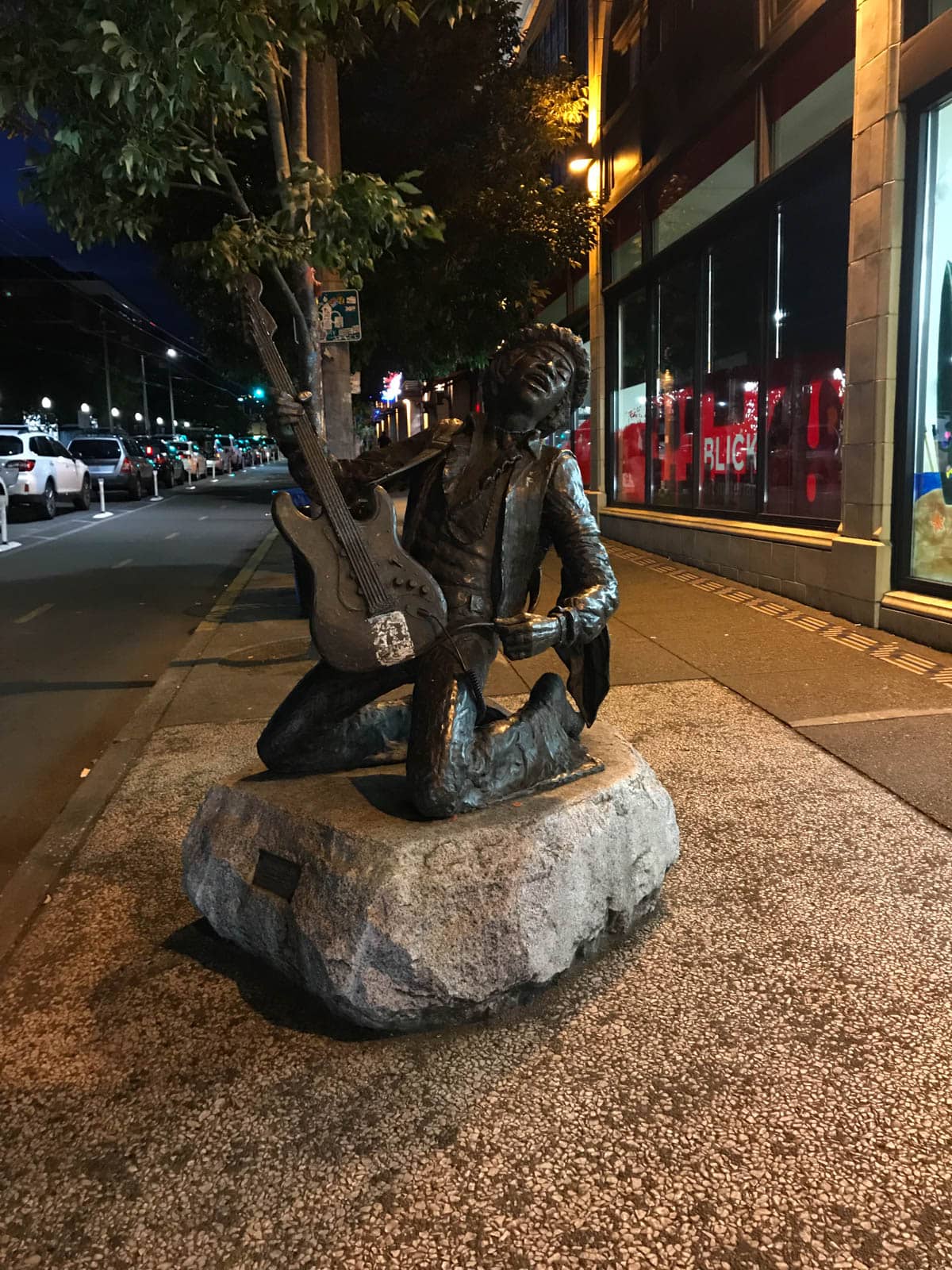 A dark bronze statue of a man playing a guitar, atop a stone with a plaque. The statue is on the sidewalk of a city, in the evening.