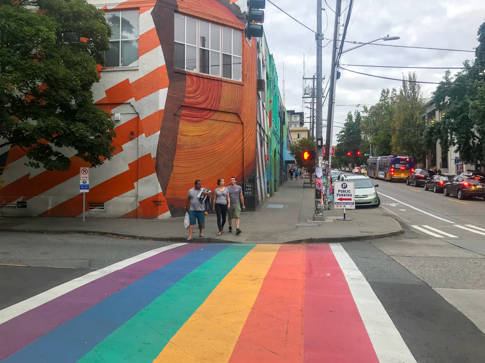 A pedestrian crossing painted in the colours of the rainbow. There are some pedestrians on the other side of the road waiting to cross, and the building behind them on the corner has a mural painted across its entire exterior