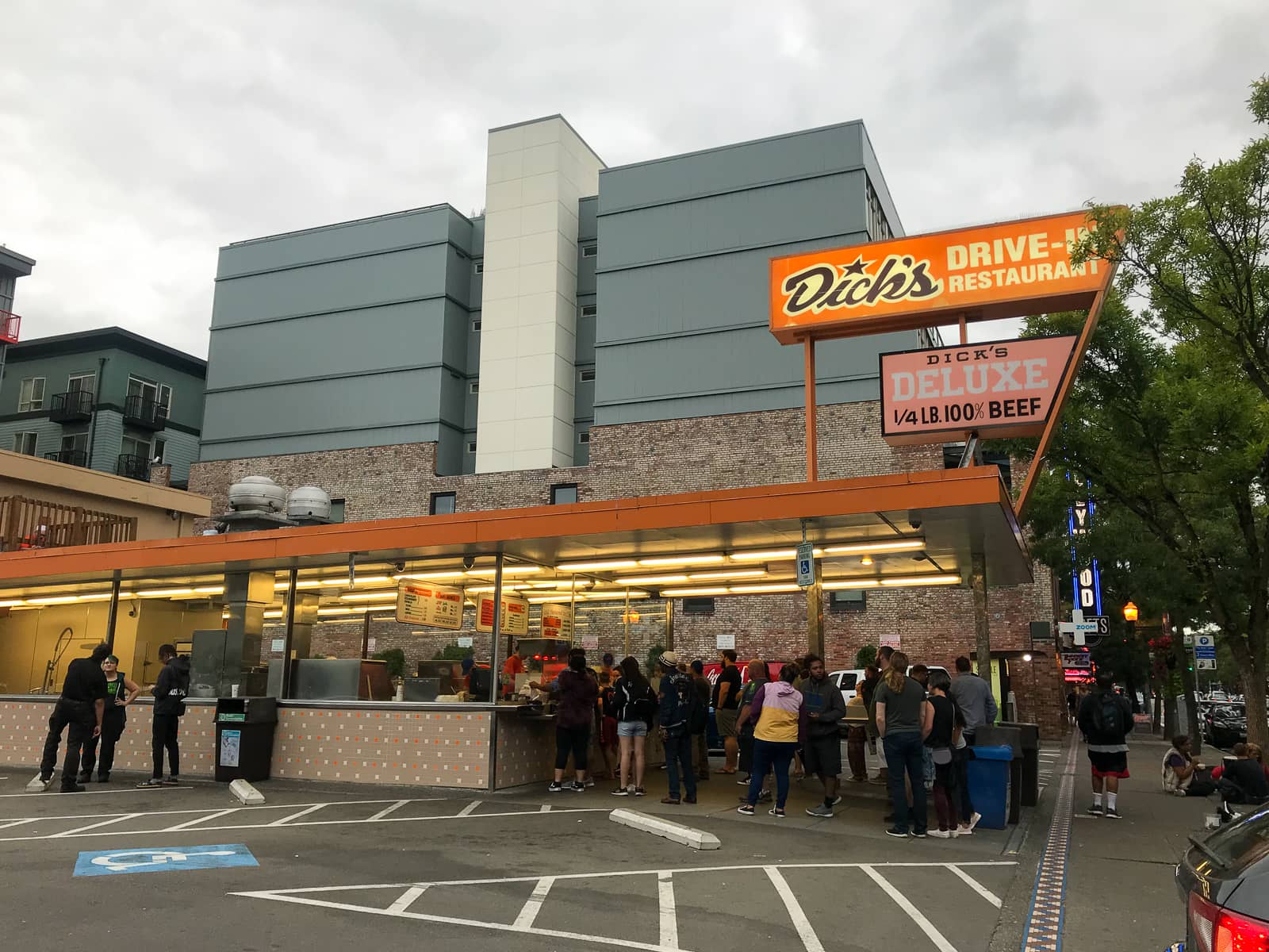 A traditional drive-in diner with a long line of people at the front. There is a sign reading “Dick’s drive-in restaurant”