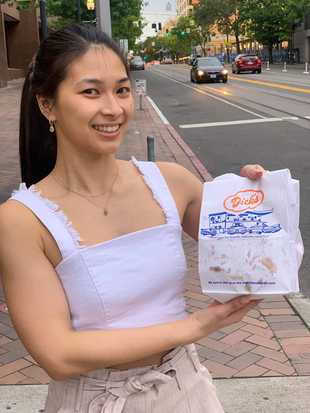 A woman with dark hair wearing a white strapless top, standing in the street holding up a paper bag reading “Dick’s”