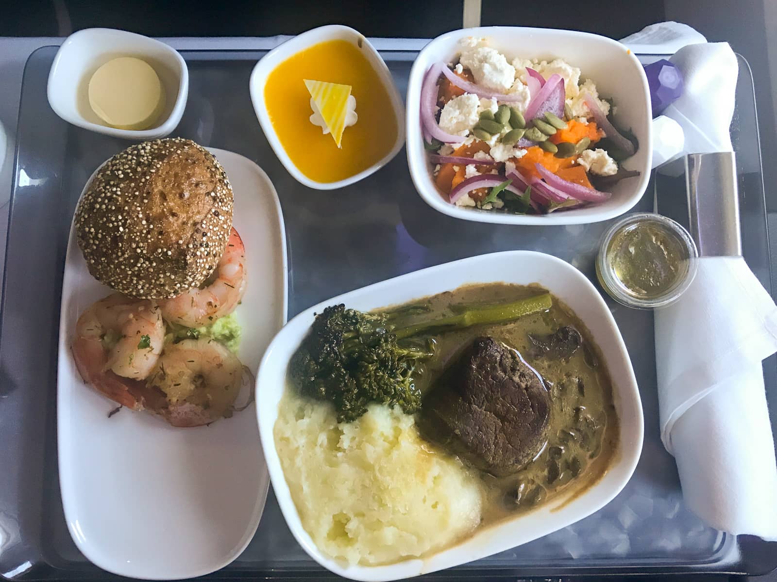 A tray of food on an airplane tray table. There is a bowl of salad; a bowl of meat, broccolini and potato; a plate of small prawns; and a small dessert