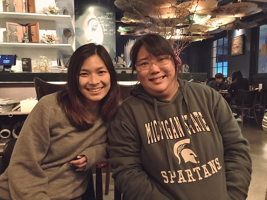 Two girls with dark hair, both wearing grey jumpers and smiling.