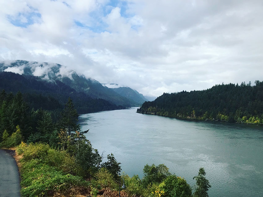 A wide river with forest terrain on either side of the river. In the distance are mountains. The sky has a lot of clouds, many of which are in the foreground.