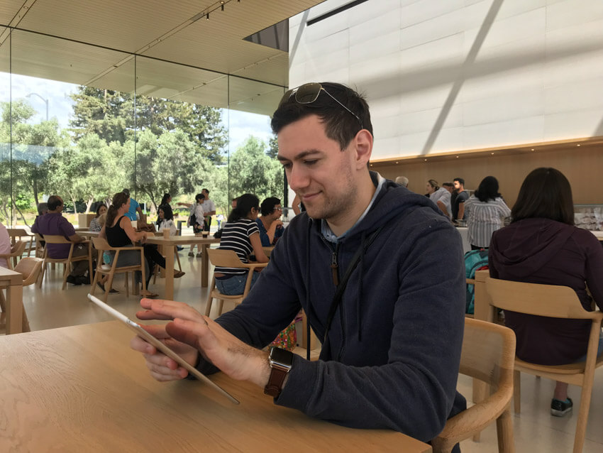 A man with dark hair, wearing a blue jacket, holding an iPad and about to press on the screen. He is sitting in a cafe with light brown furniture and full-size windows showing trees outside.