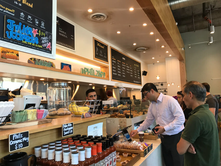 The counter at the inside of a cafe. A man is paying for his purchase, and in the foreground are some samples of food as well as bottles of hot sauce for sale. There are blackboards above the counter showing the menu.