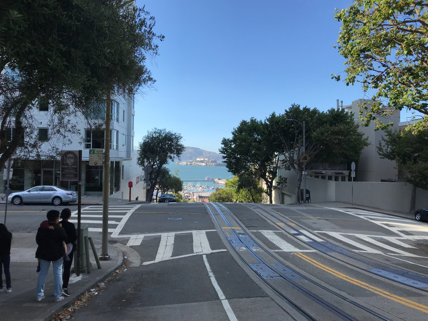 The intersection of a road, on top of a steep hill, showing water and a blue sky in the distance.