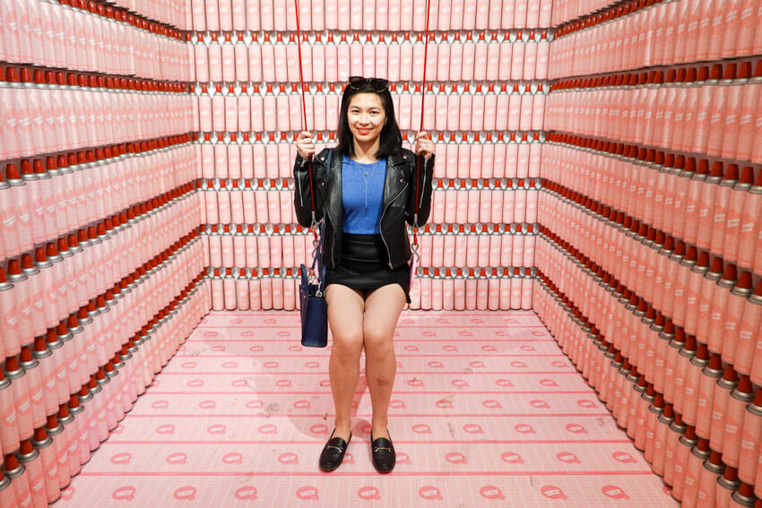 A woman wearing a black skirt and jacket sitting on a swing in a small cubicle with pink spray cans lining the walls