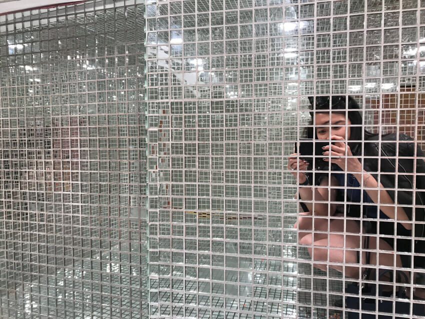 A woman taking a photo in a room with entirely mirrored surfaces of small reflective square tiles. The woman is crouched down with a jacket draped over her shoulders.