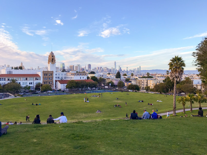 A view of a city from park on top of a hill. The park has very green grass, and it’s late afternoon and the city in the distance looks faded. The sky is light blue with some scattered clouds.