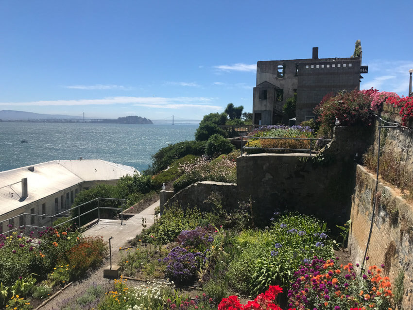 A garden on an island, bordered by a stone wall in a step-like pattern. In the background is a bridge on the water.