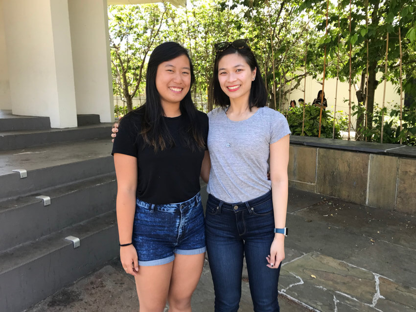 Two women joined at the hip, one wearing a black shirt and the other wearing a grey shirt. Both have dark hair and are smiling.