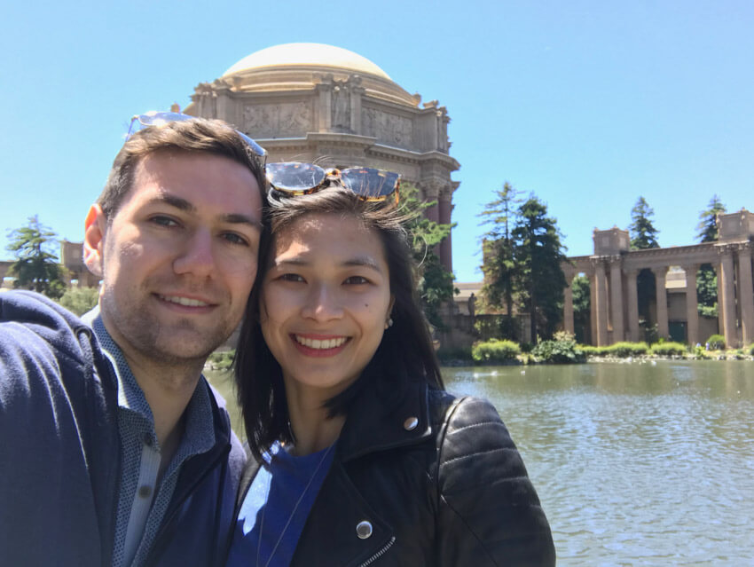 A man and woman smiling, in front of a lake with an old dome-shaped building on the other side of the lake.