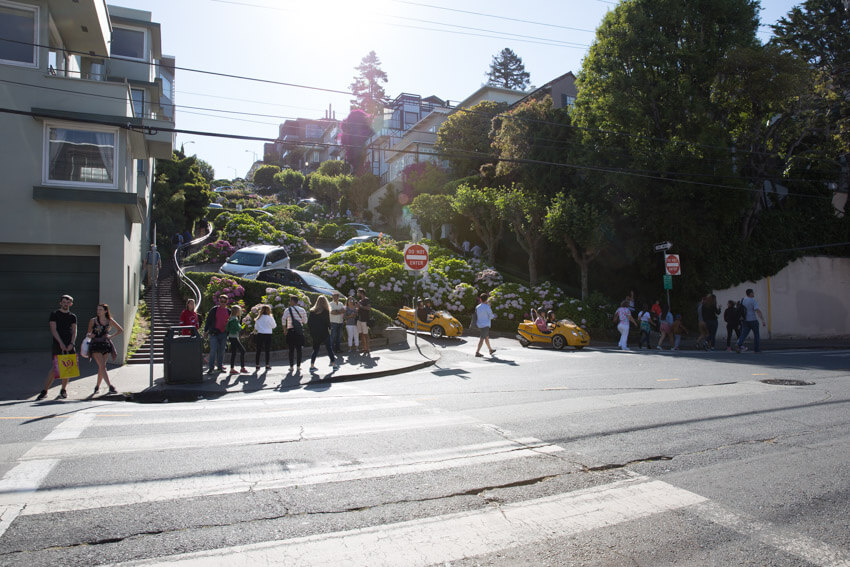 A steep street in a zig-zag shape, going upwards. Cars fill the street, and bright green trees and pink flowers fill in the median areas. A staircase is at the side of the street. A crosswalk heading towards the zig-zag street is in the foreground.