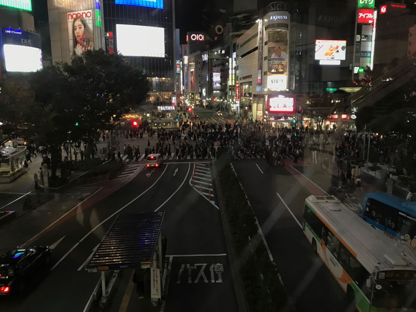 A view of a street intersection through a glass window at night. A very large amount of people cover the areas of the intersection where crossings are marked. Some buses can be seen on the right, moving in the direction of where the photo was taken from. Multi-storey buildings with neon signs of store logos are in the background. Some reflections are visible in the foreground