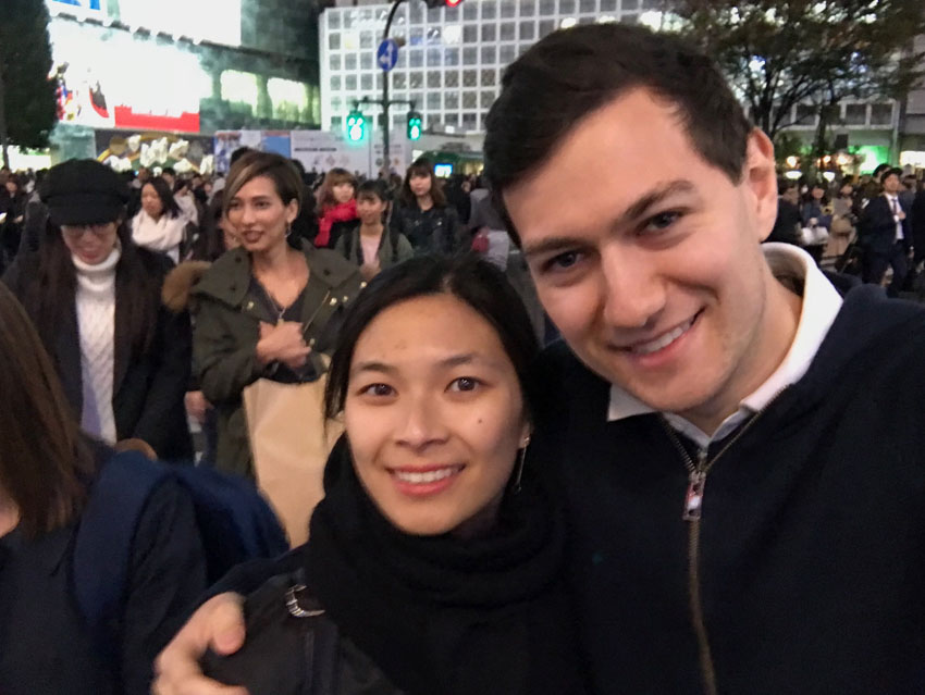 A boy and a girl smiling, wearing dark clothes. In the background are a lot of people walking.