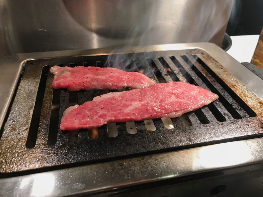 Two slices of red raw meat on a hot grill, with some smoke rising from them.