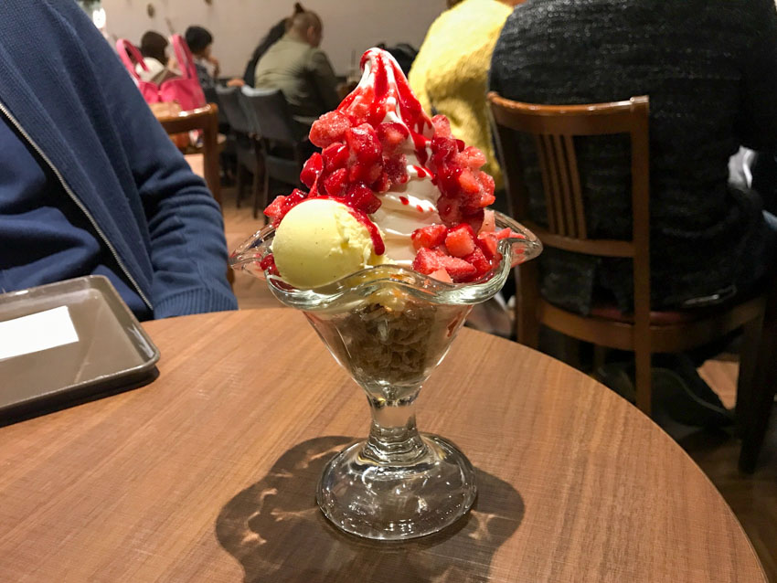 A parfait sitting on a round wooden table in a cafe setting. The parfait is presented in a glass that has curved edges. Flakes fill the bottom of the glass, it is topped with white cream, a round ball of ice cream, chopped strawberries and red strawberry sauce.