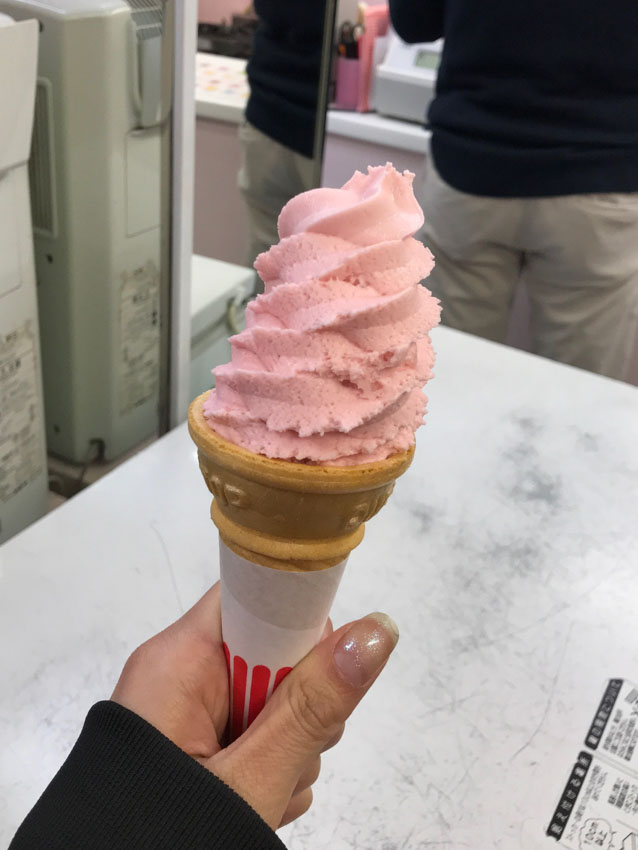 An ice cream cone in someone’s hand. The ice cream is light pink and is served soft-serve style.