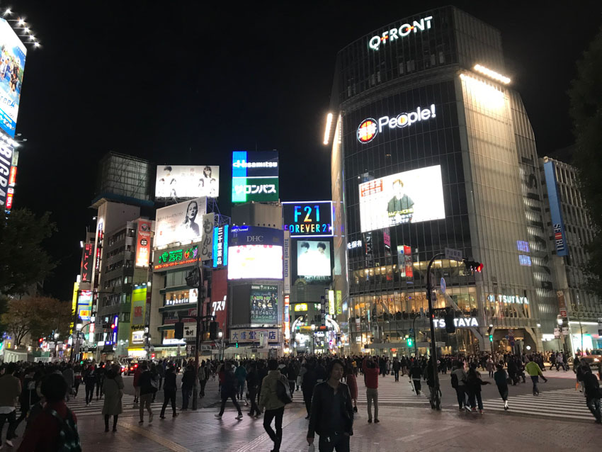 A busy intersection at night time with a lot of pedestrian traffic. Three crossings span out straight in front, at 10 o’clock, and 2 o’oclock. High-rise shopping buildings are ahead up ahead.