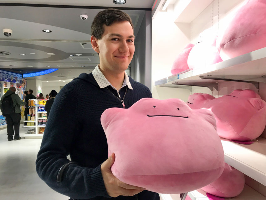 A man with dark hair wearing a navy jacket, smiling, holding a big pink blob-shaped plushie toy with a smiling face.
