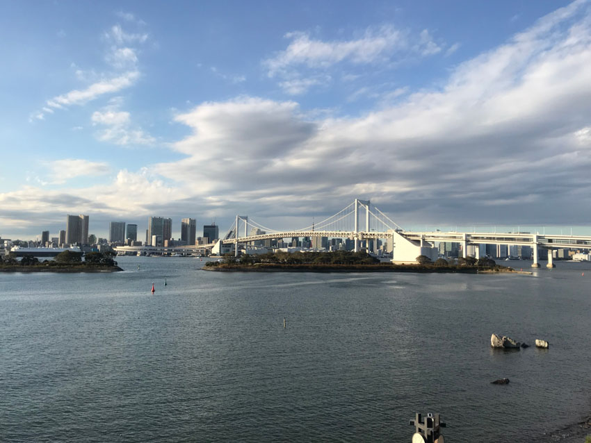 A view of a bridge on the water. There is an island of trees in the foreground. The sky is light blue with many white clouds.