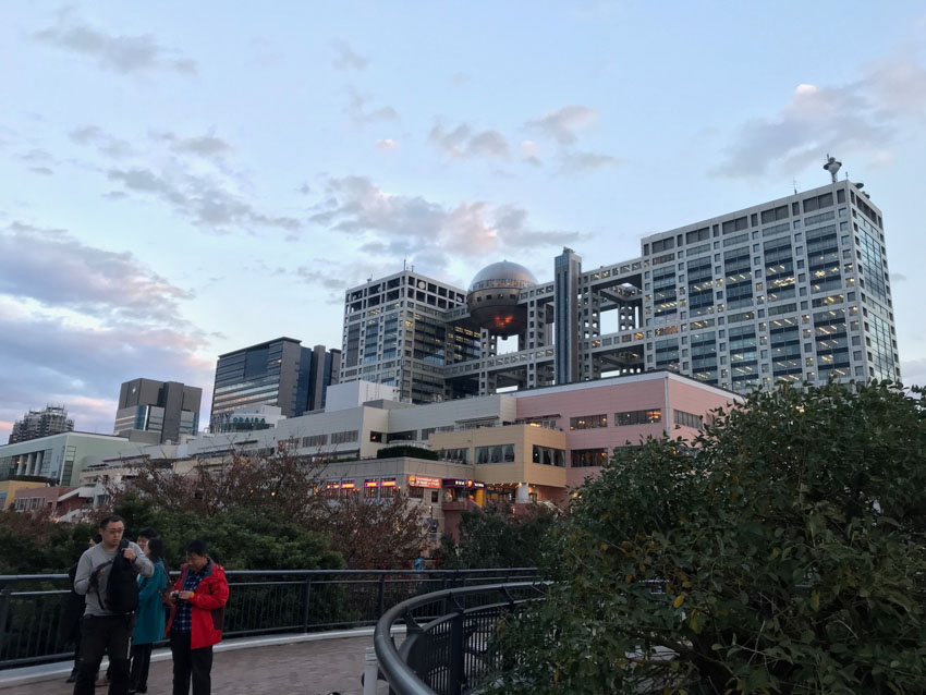 An elevated path with a black railing on either side curves to the right. There are the tops of trees on either side of the path. In the distance are high-rise buildings, one of which has a spherical feature on it. It is dusk and the blue sky looks dim.