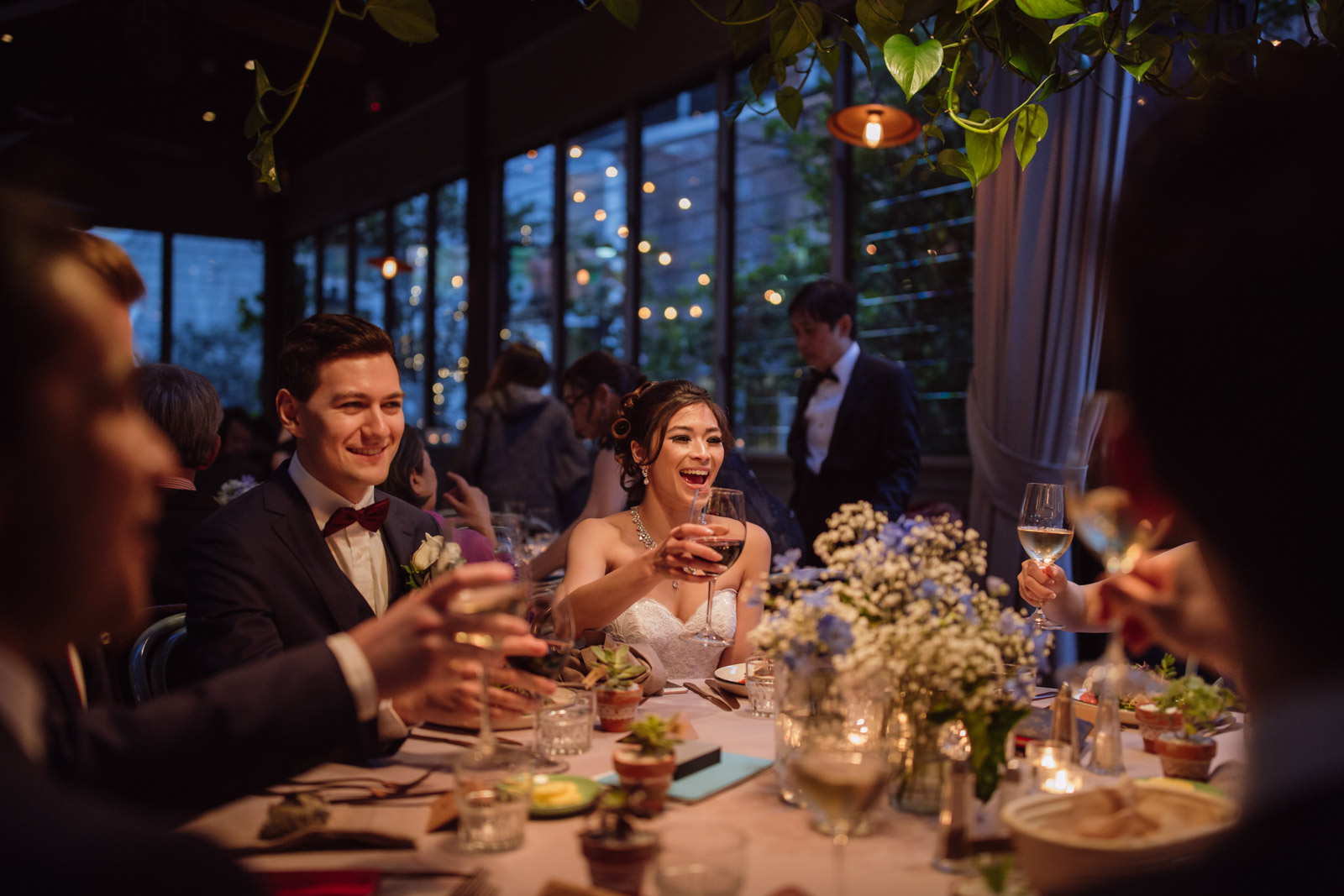 A man and woman at a round table, in an evening setting and dim lighting, smiling, holding glasses of wine up for a toast. Some other people are at the table but not fully in view.