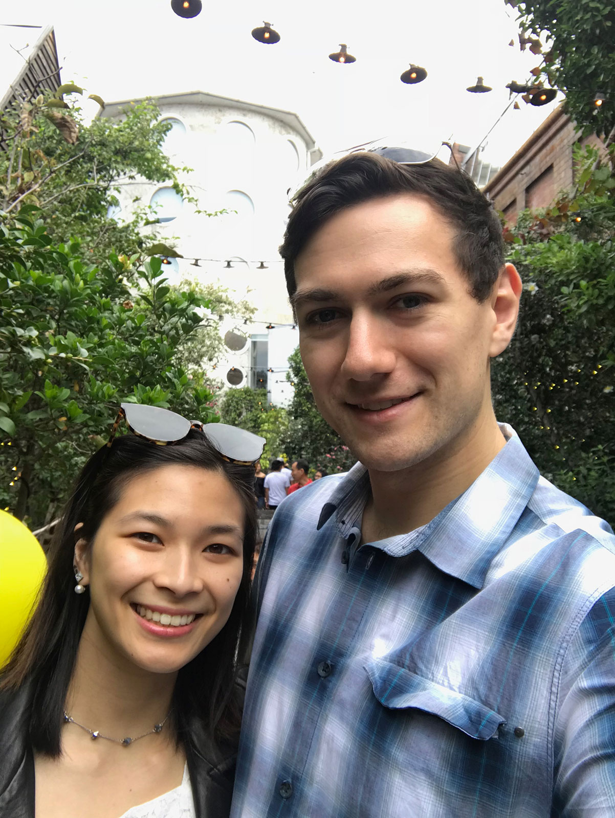 A man and woman smiling. They both have sunglasses on top of their heads. The man is wearing a blue checkered shirt and the woman is wearing a white top, black jacket and thin choker necklace. In the background are a lot of green leaves from trees and plants.