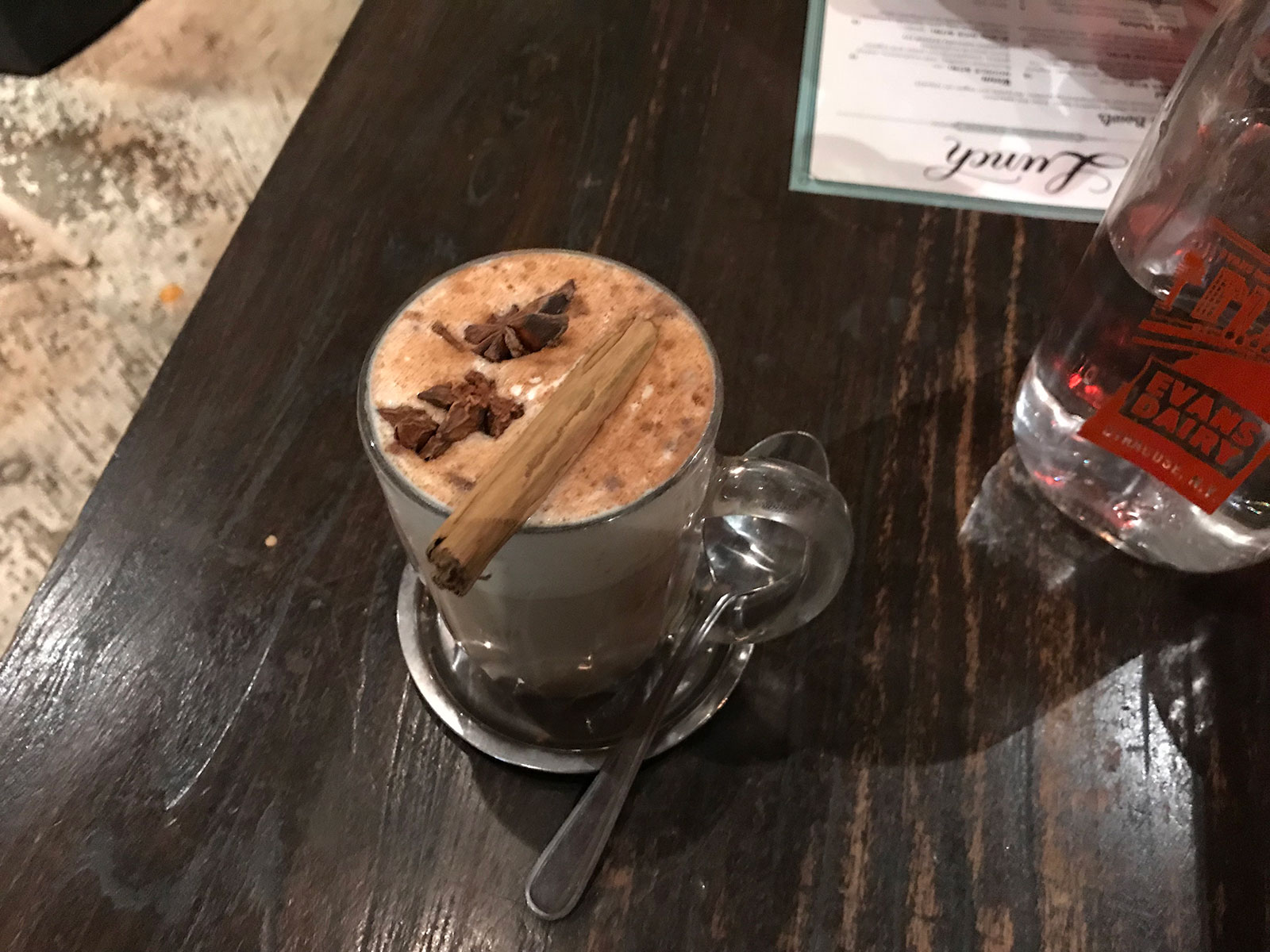 A glass with a handle, filled with a foamy drink, sitting on a table, and a silver spoon through the handle of the glass. The drink has a cinnamon stick and a few pieces of star anise floating in it.