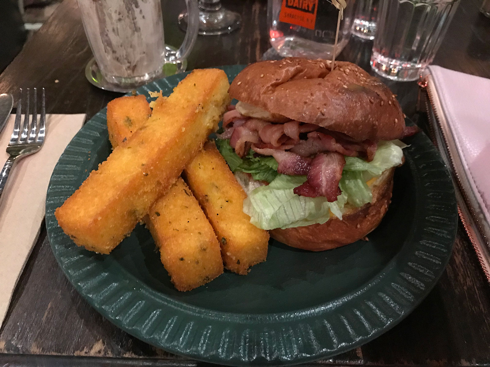 Three large rectangular yellow polenta chips, and a burger filled with visible lettuce and bacon, served on a dark green plate on a table.