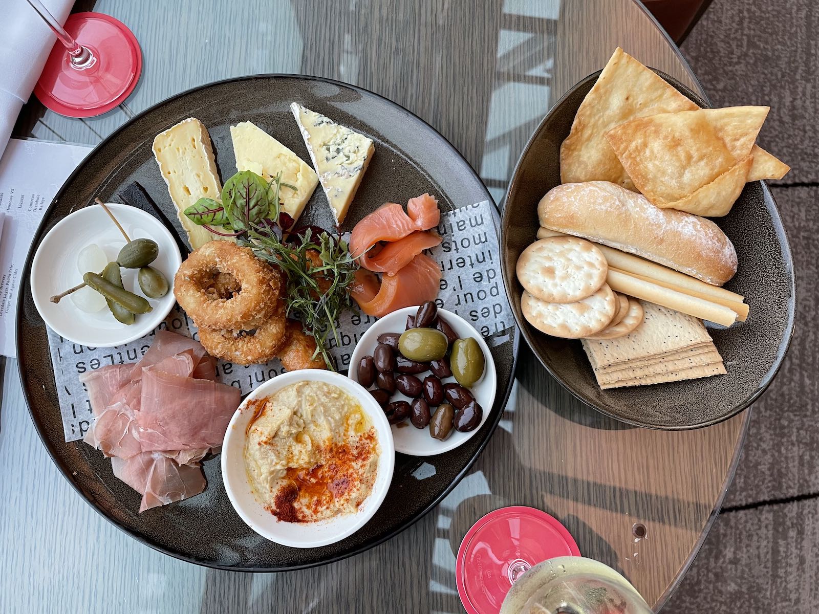 A top-down view of a cheese and meat platter with some olives and pickles, and a small bowl of bread and crackers sitting on a glass table