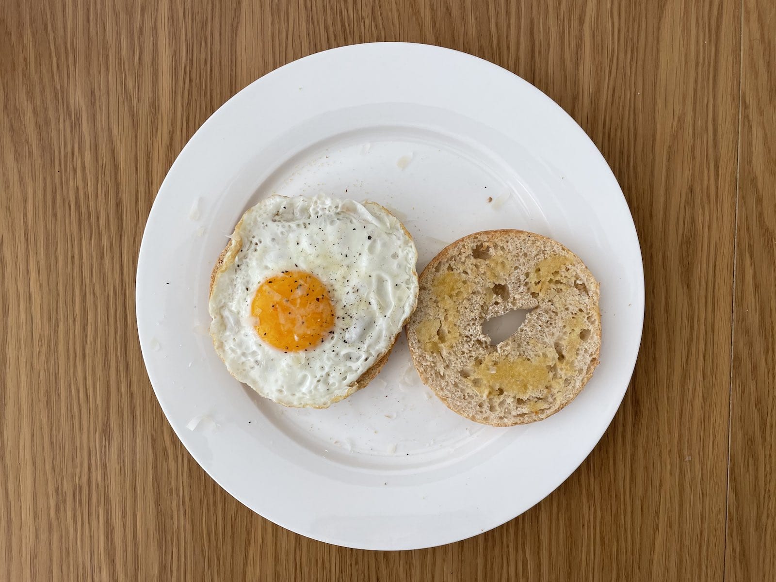 An open bagel served with a fried egg on top of one half, sitting on a white plate on a wooden table